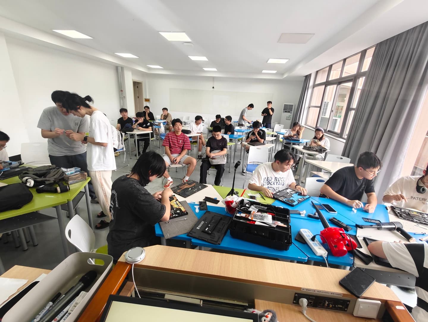 电脑医院在借用教室里集中开展维修活动,同学们分散在各张桌子前动手